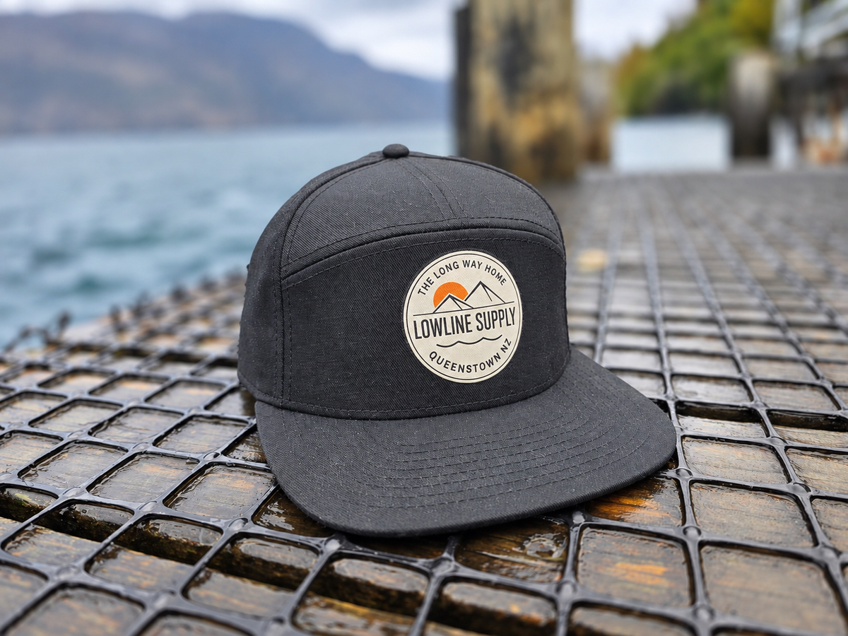 A dark charcoal gray snapback cap with a circular embroidered 'Lowline Supply' logo patch, resting on a wet metal grate with a blurred lake and mountains in the background.
