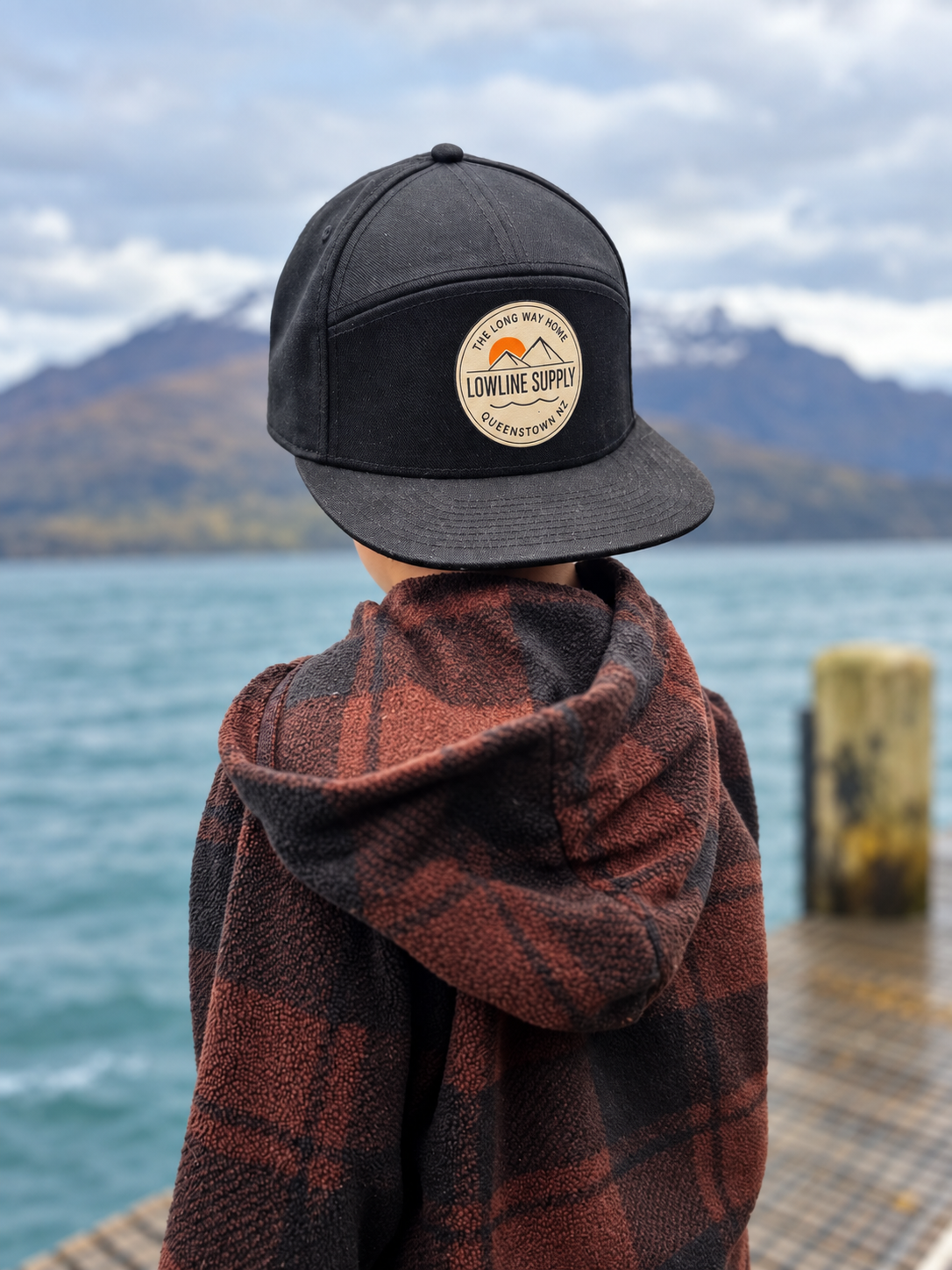 A person seen from behind wearing a black flat-brim cap with a circular 'Lowline Supply' patch, a rust and black plaid hooded blanket, and standing on a dock with water and mountains in the background.
