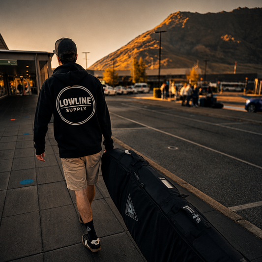 Person wearing a 'Lowline Supply' hoodie walking with a surfboard case to the airport with mountains in the background.