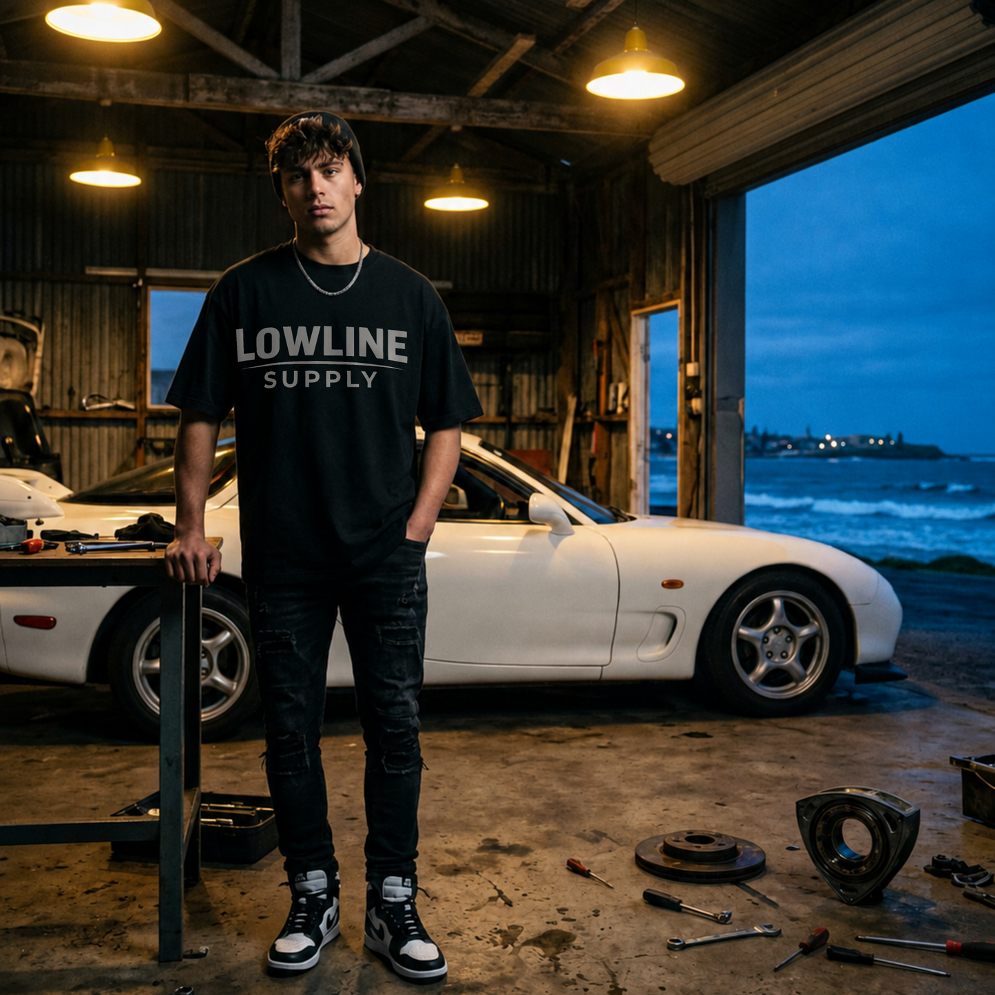 Person wearing a 'LOWLINE SUPPLY' t-shirt standing in a garage with a Mazda rx7 car and ocean view.