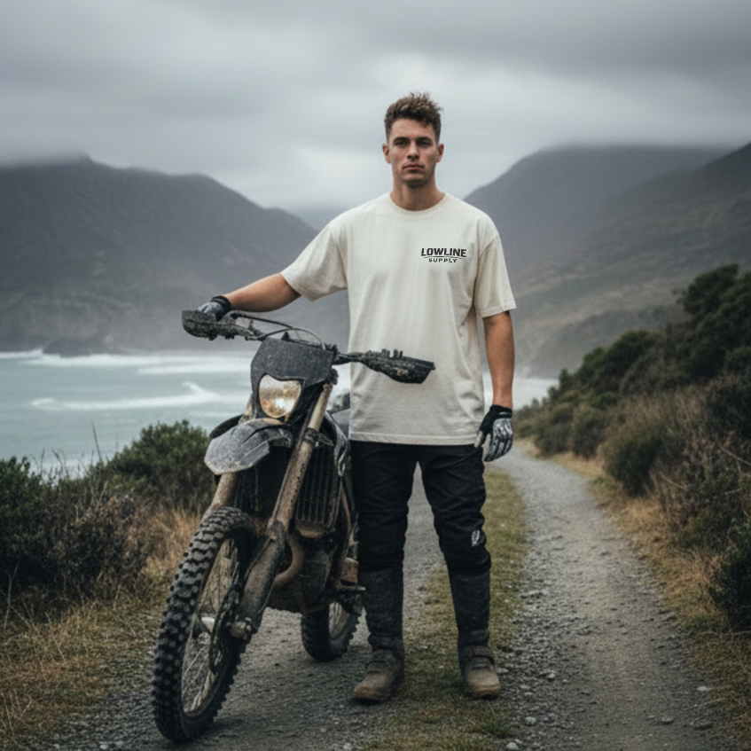 Man standing next to a dirt bike on a scenic trail with mountains and water in the background