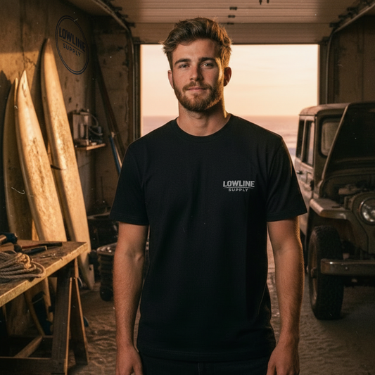 Man standing in a garage with a jeep and surfboards, wearing a black t-shirt with a logo.